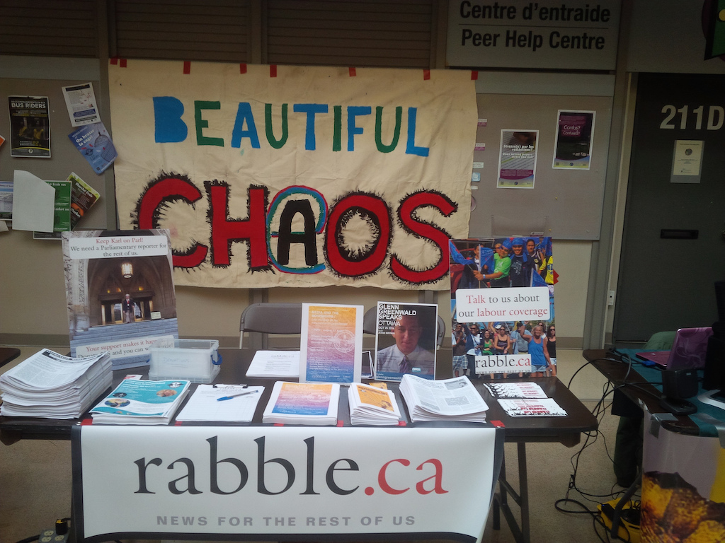 A table with a rabble.ca banner, covered in pamphlets with a large hand-painted sign behind which reads 'Beautiful CHAOS'. 