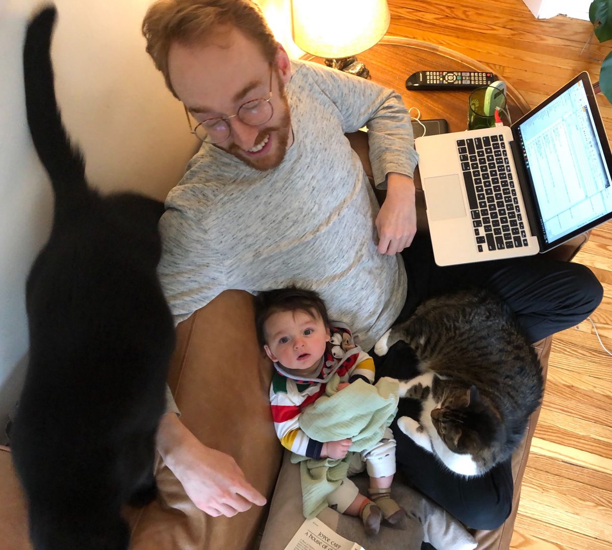 Fraser sits on the couch with his laptop, his one year old son and two cats.