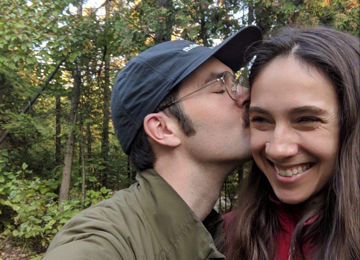Claudia and John in the woods. Claudia smiles as John kisses her on the side of her head.